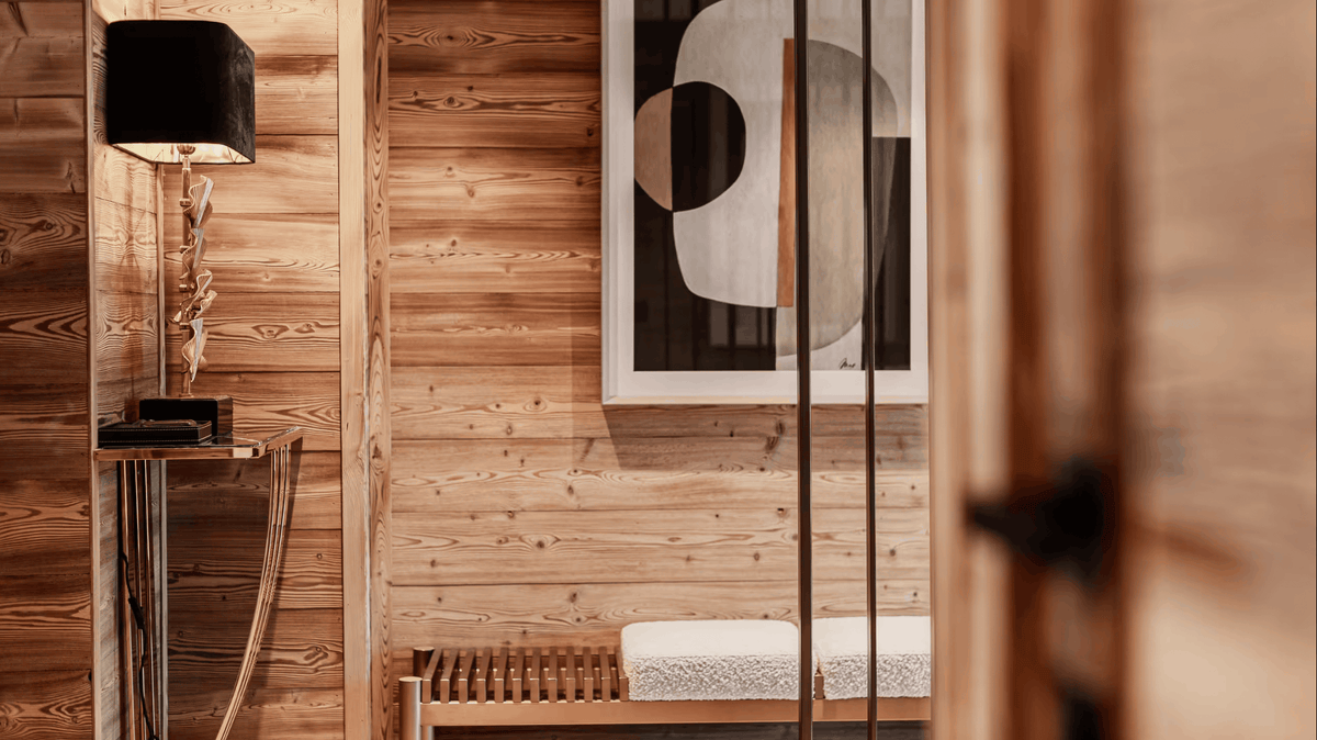 A warm-toned interior scene with wood-paneled walls, a black floor lamp on a side table, a framed abstract black-and-white artwork, and a wooden bench with a light cushion in the foreground.