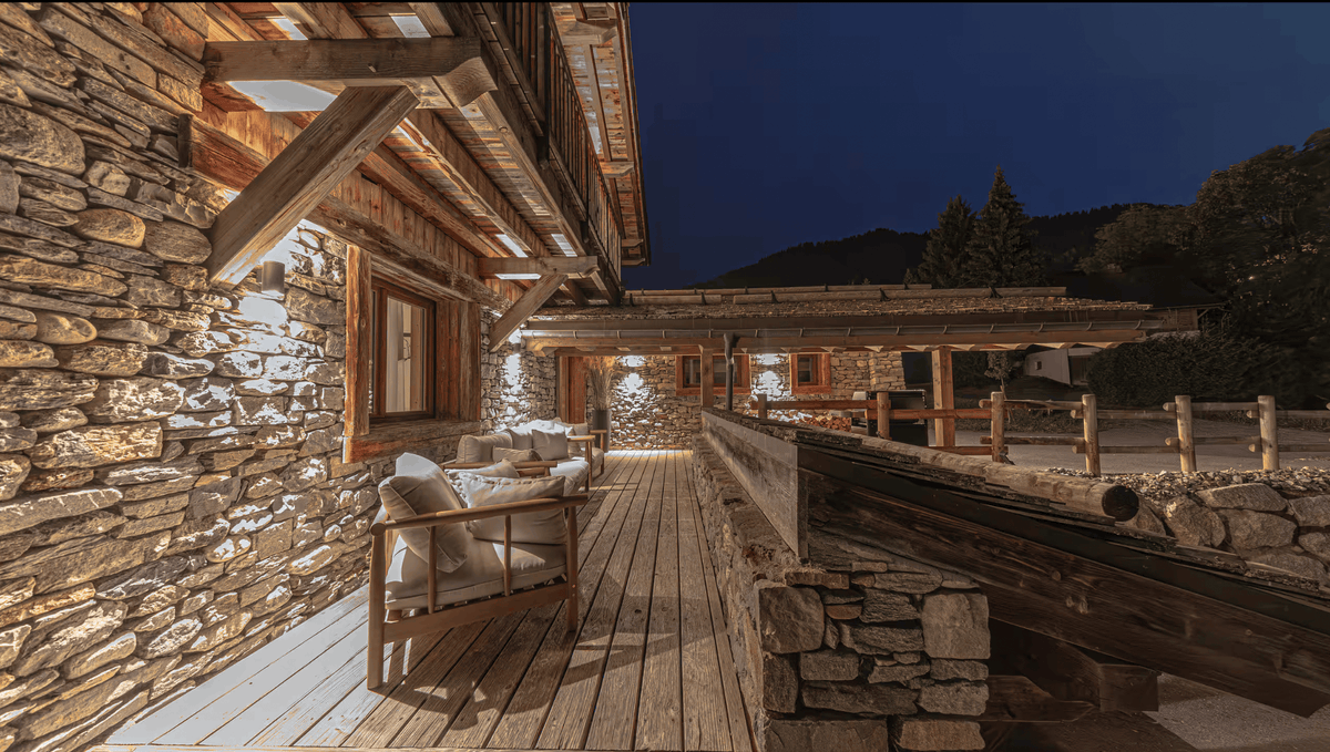A rustic wooden chalet patio at night with stone walls, warm outdoor lighting, a wooden chair with cushions, and a covered deck leading toward the building.