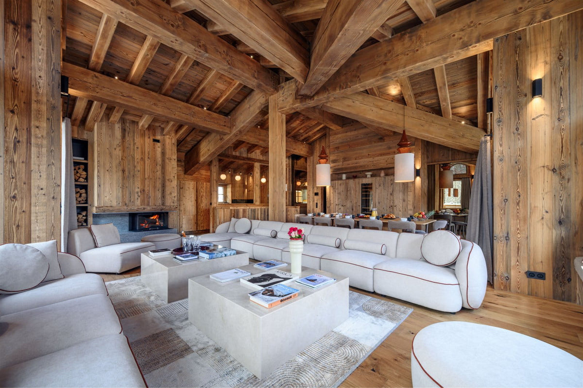 A luxurious modern living room with a large white sectional sofa, low concrete coffee tables, a fireplace, and a high wooden ceiling with exposed beams.
