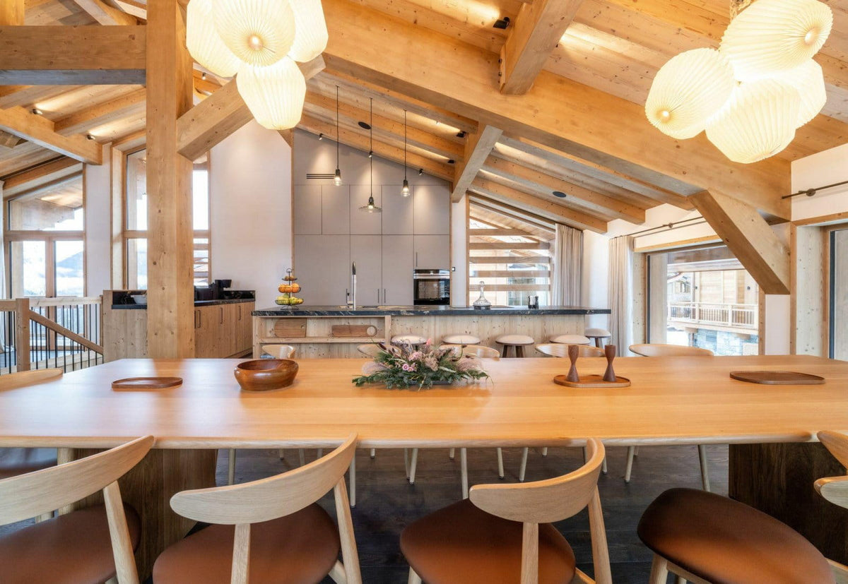 A modern ski resort chalet dining area featuring a large wooden dining table with light wood chairs, under a high wooden ceiling with exposed beams and decorative pendant lights.