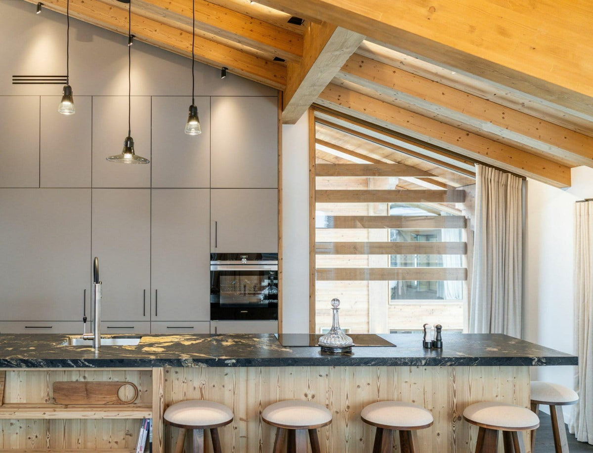 A modern kitchen interior with light wood cabinetry, a dark stone countertop, pendant lights, exposed wooden ceiling beams, and four bar stools at the island.