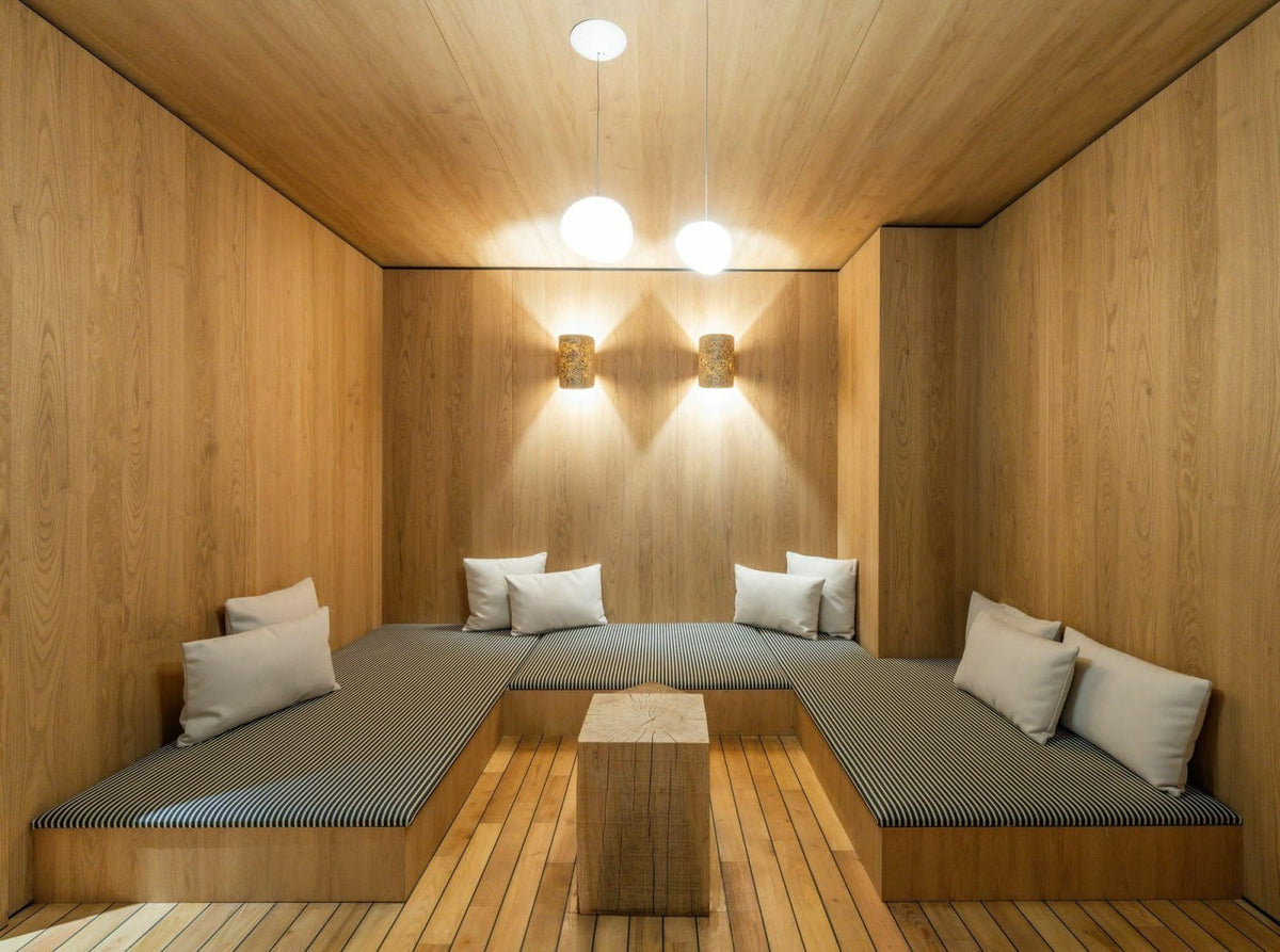 A cozy wooden sauna or relaxation room with built-in cushioned seating, light wood walls and ceiling, gray ribbed seat cushions, white pillows, and two hanging globe lights above a small wooden table.
