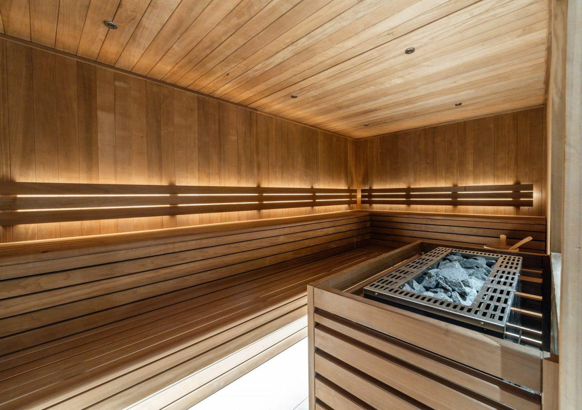 A warm-toned wooden sauna interior with tiered seating, built-in lighting, and a metal firebox with glowing coals in the foreground.