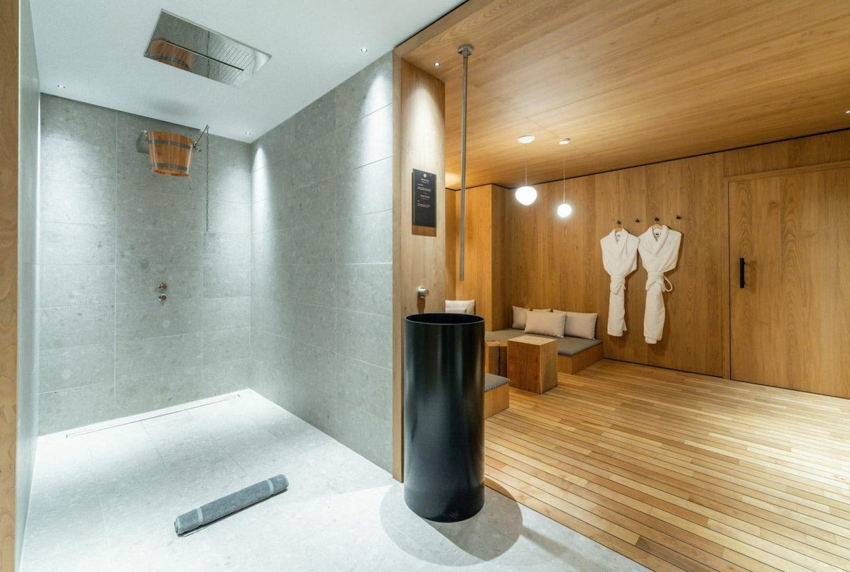 A luxury spa bathroom interior featuring a modern walk-in shower with gray stone walls, a matte black cylindrical pedestal sink, light wood flooring, and a warm wood-paneled relaxation area with white robes hanging on hooks.
