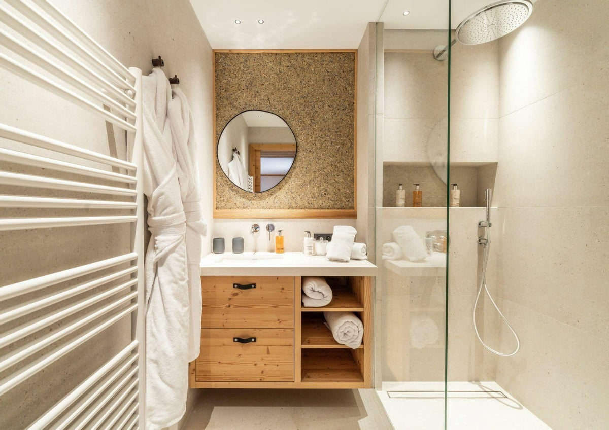 A modern bathroom vanity area with a light wood cabinet, white countertop, round mirror, and open shelving, next to a glass shower enclosure and a white towel rack.