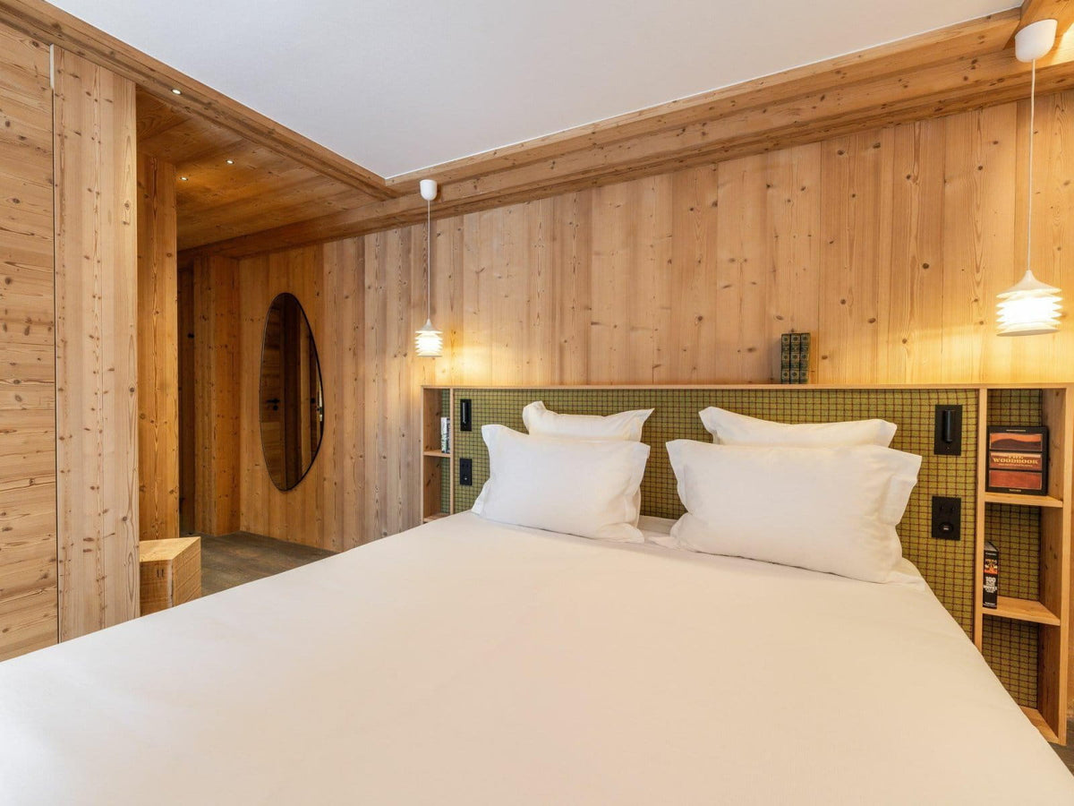 A modern bedroom with a large bed dressed in white linens, set against light wood-paneled walls with built-in shelving and pendant lights.