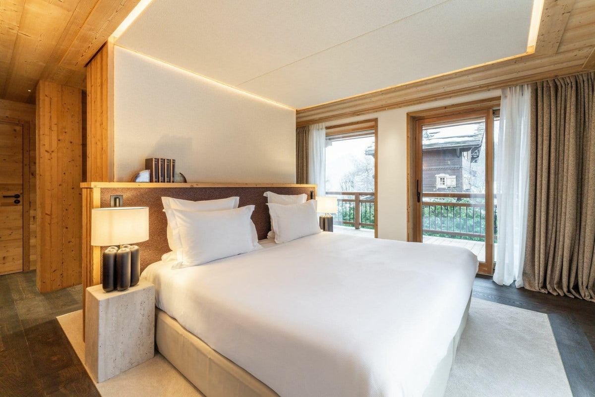 A modern luxury bedroom featuring a large bed with crisp white linens, a dark wood headboard, and built-in bedside lamps. The room has warm wood-paneled walls and ceiling, large windows with white and beige curtains, and a balcony view.