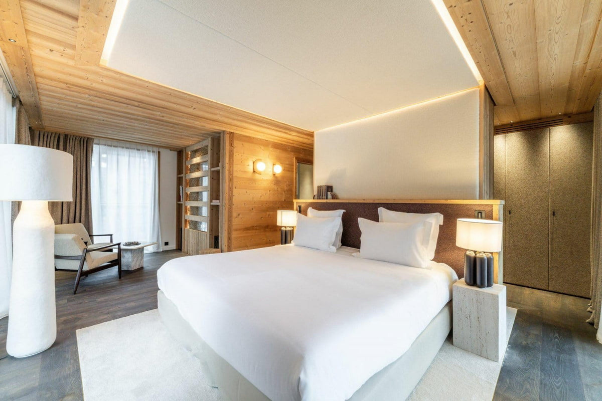 A modern luxury bedroom featuring a large bed with crisp white linens, a dark wood headboard, warm wood-paneled walls and ceiling, bedside tables with lit lamps, and a light rug on a dark wood floor.