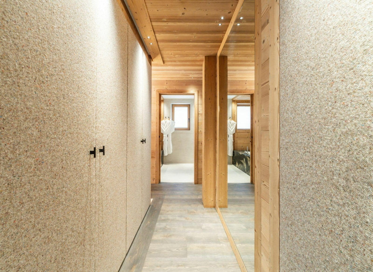 A modern interior hallway with textured beige walls, light wood ceiling and columns, and gray stone-look flooring, leading toward open rooms with white robes visible in the distance.