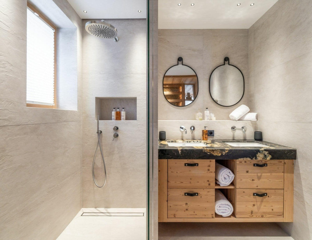 Modern bathroom interior with a walk-in shower area on the left and a vanity with two sinks, round mirrors, and a wood cabinet on the right. The space has light stone walls and a dark marble countertop.