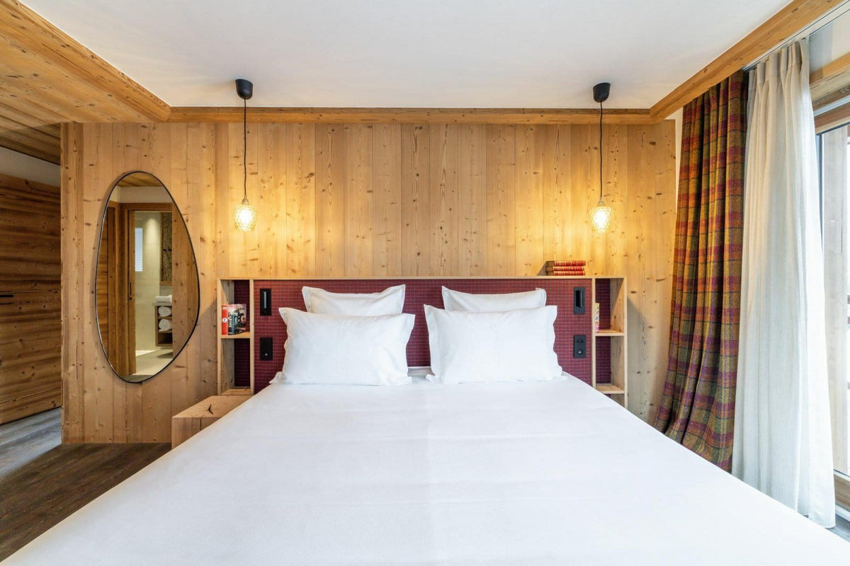 A cozy bedroom interior with a neatly made bed in the foreground, light wood paneled walls, two hanging pendant lights, a round wall mirror, and plaid curtains by a window.