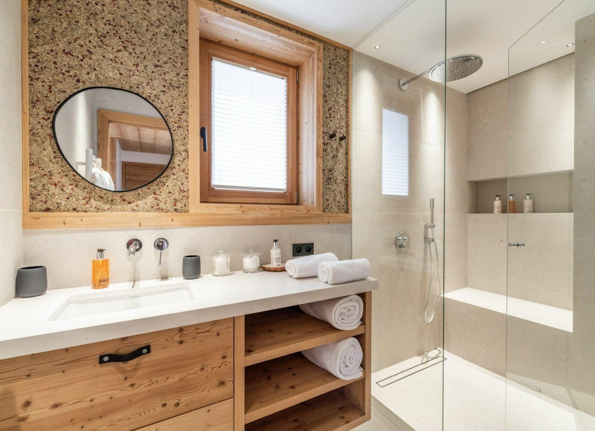 Modern bathroom interior with a wooden vanity, white countertop, round mirror, open shelving with rolled towels, and a glass shower enclosure.