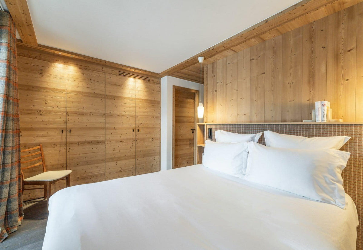 A modern bedroom with a large bed dressed in white linens, set against a light wood-paneled wall with warm ambient lighting. A matching wood wardrobe is visible on the left, along with a chair and plaid curtain.
