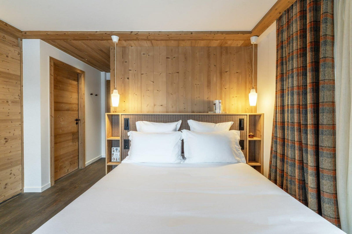 A modern bedroom featuring a neatly made bed with white linens, set against a light wood-paneled wall with built-in shelving. Warm pendant lights glow above the bed, and a plaid curtain hangs on the right side.