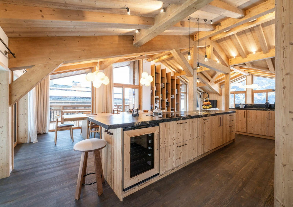 A modern kitchen island with light wood cabinetry and dark countertops in a rustic alpine-style chalet with exposed wooden beams and large windows.