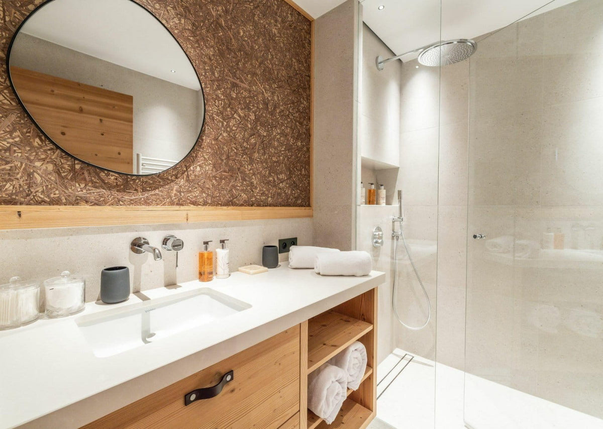 Modern bathroom vanity area with a white countertop, wood cabinetry, a large round wall mirror, and a glass shower enclosure.