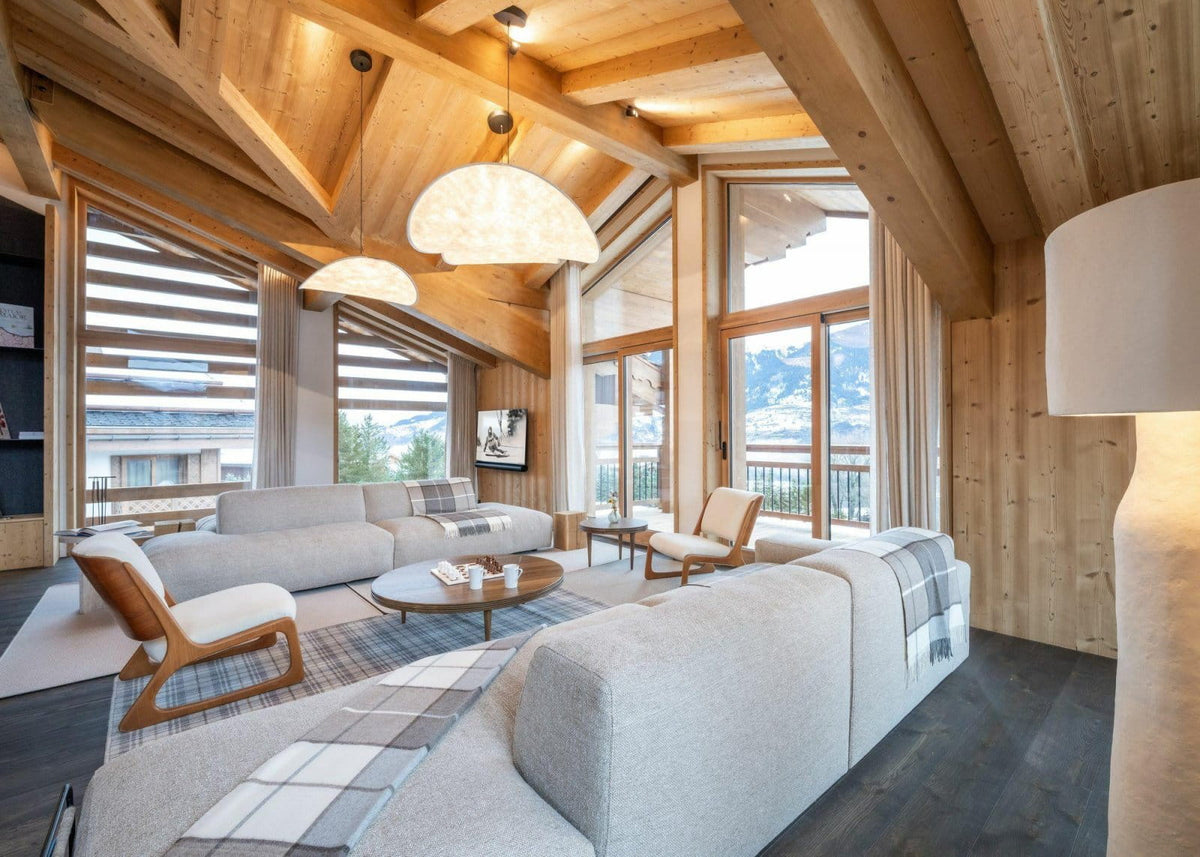 A modern ski chalet living room featuring light gray upholstered sectional sofas, wooden armchairs, a round coffee table, and large windows with mountain views.