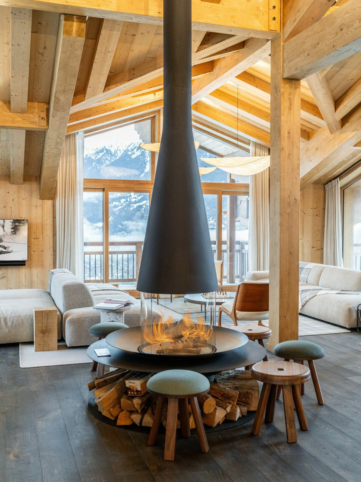 A modern wood-burning fireplace with a tall black chimney hood, surrounded by stacked firewood and four round green stools, set in a rustic wooden chalet living room with a mountain view.