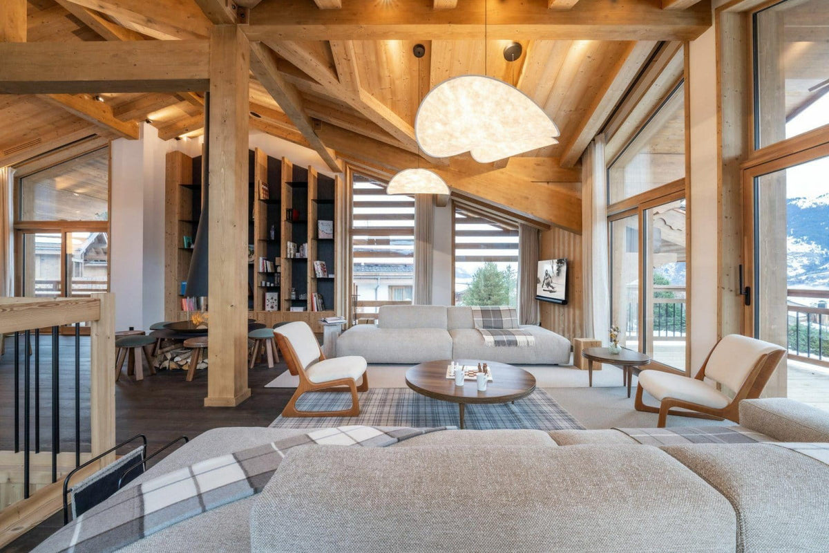 A bright, modern mountain chalet living room with high wooden beams, large windows, and a light beige sectional sofa in the foreground. The room features two matching armchairs, a round wooden coffee table, built-in dark wood bookshelves, and a view of snowy mountains outside.