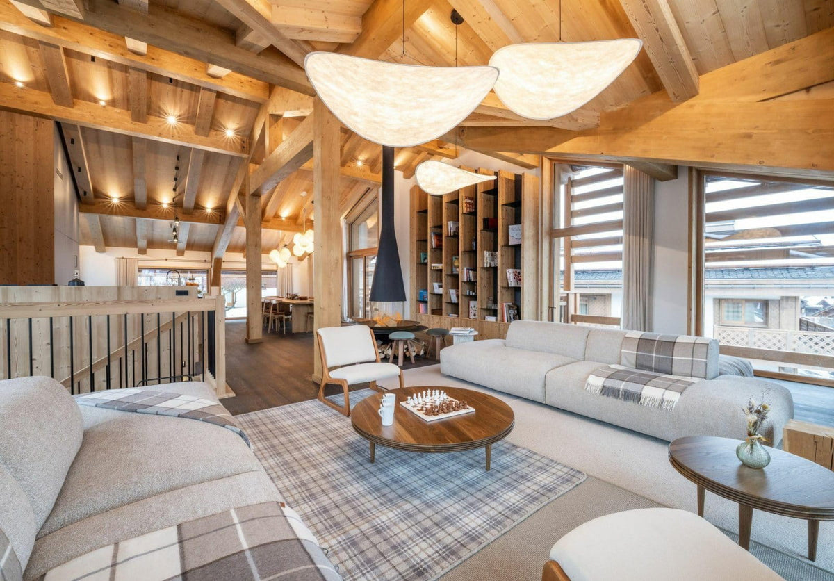A bright, modern chalet-style living room with a large white sectional sofa, round wooden coffee table, plaid rug, exposed wood ceiling beams, and large windows.