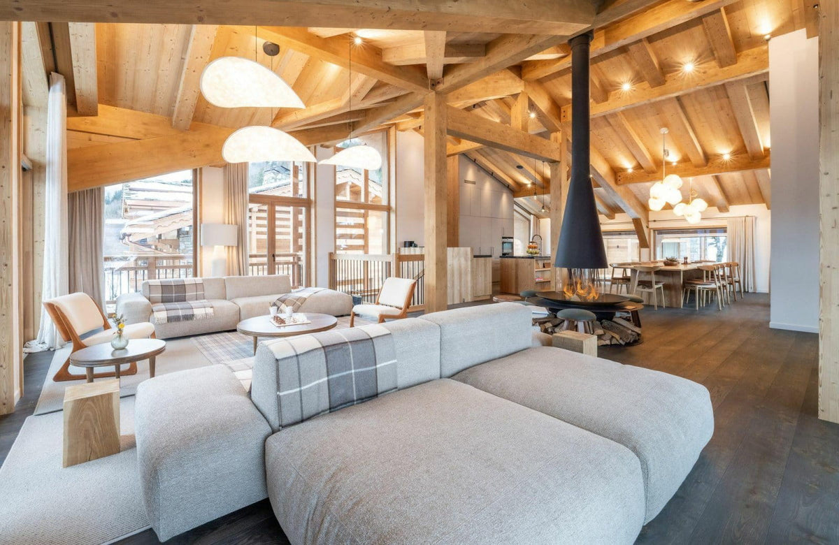 A modern light gray modular sectional sofa in a spacious, rustic chalet-style living room with exposed wooden ceiling beams, a hanging black fireplace chimney, and large windows.