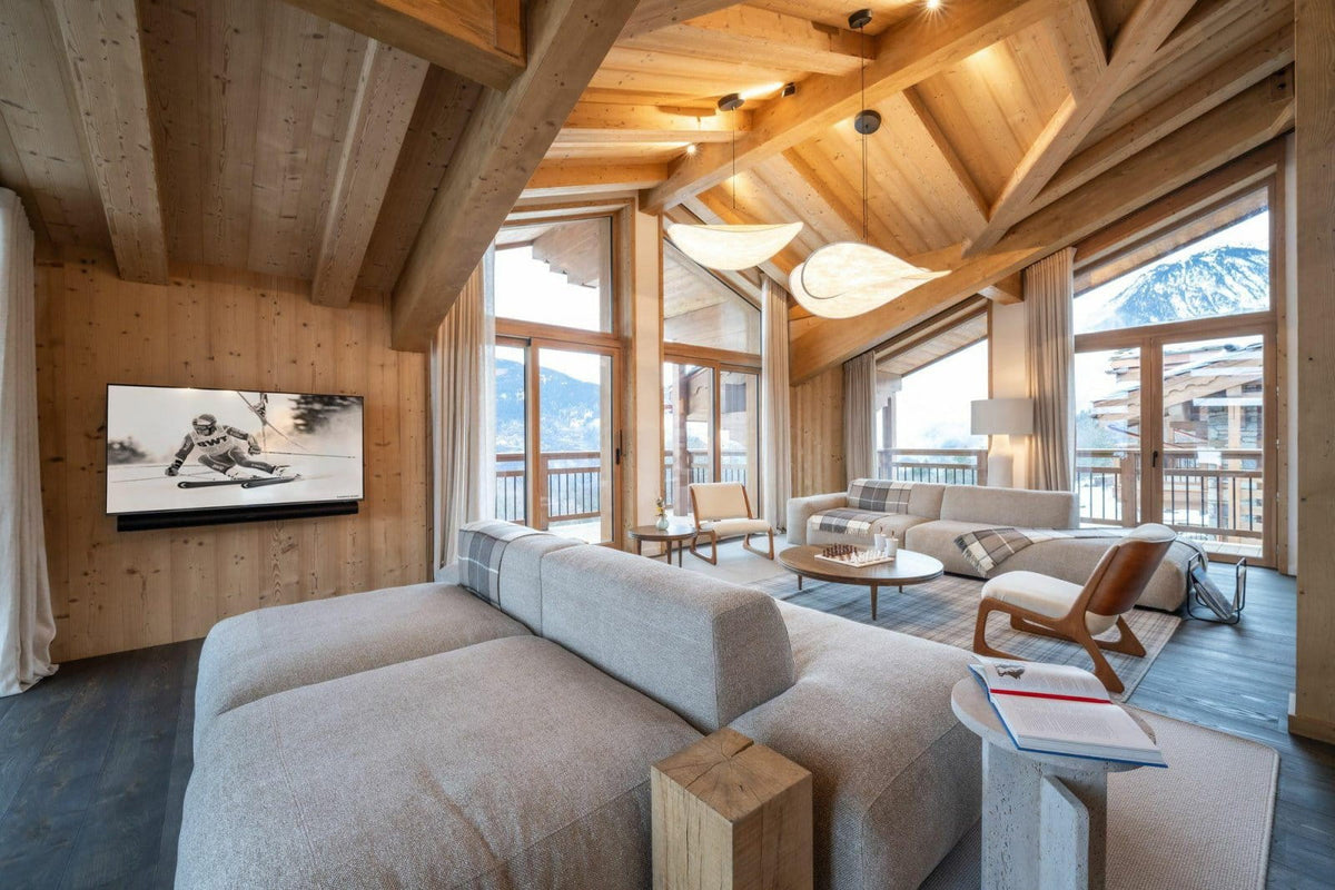 A modern ski chalet living room with a large light gray sectional sofa in the foreground, wood-paneled walls and ceiling, large windows, and mountain views outside.