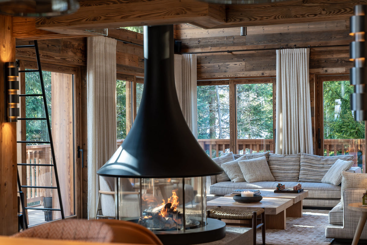 A modern black conical fireplace with a visible fire, set in a rustic wooden cabin living room with a sofa, coffee tables, and large windows looking out to trees.