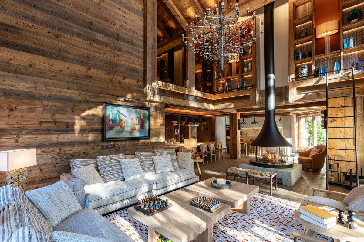 A high-ceilinged rustic lodge living room with wood-paneled walls, a large light gray sectional sofa, a patterned rug, a black freestanding fireplace chimney, built-in wooden shelves, and a chandelier overhead.