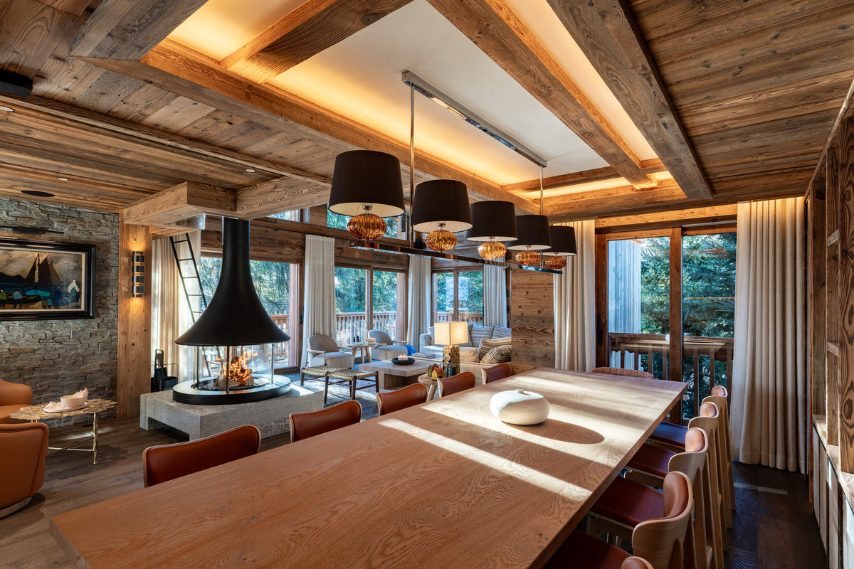 A luxury modern chalet interior featuring a long wooden dining table with red upholstered chairs, a black chimney pot fireplace, stone walls, and a rustic wood-beamed ceiling.
