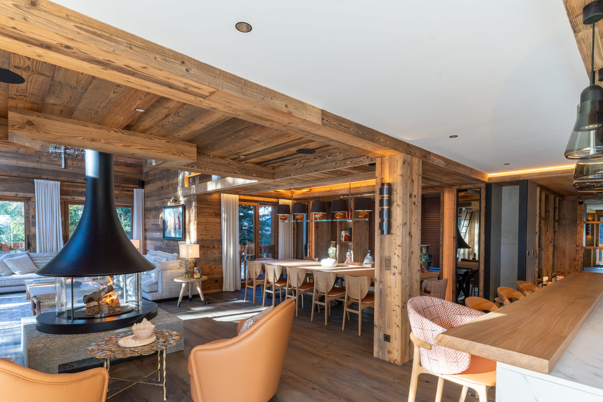 A luxurious modern chalet interior featuring a large black conical fireplace, rustic wooden beams and floors, a dining area with light wood chairs, and a kitchen island with pink upholstered bar stools.