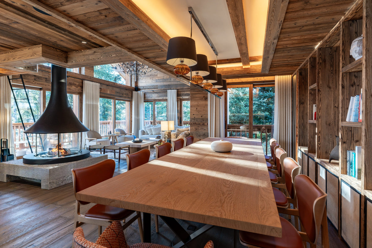 A long wooden dining table with reddish-brown upholstered chairs in a rustic lodge-style dining room with exposed wood ceiling beams, large windows, and a fireplace.