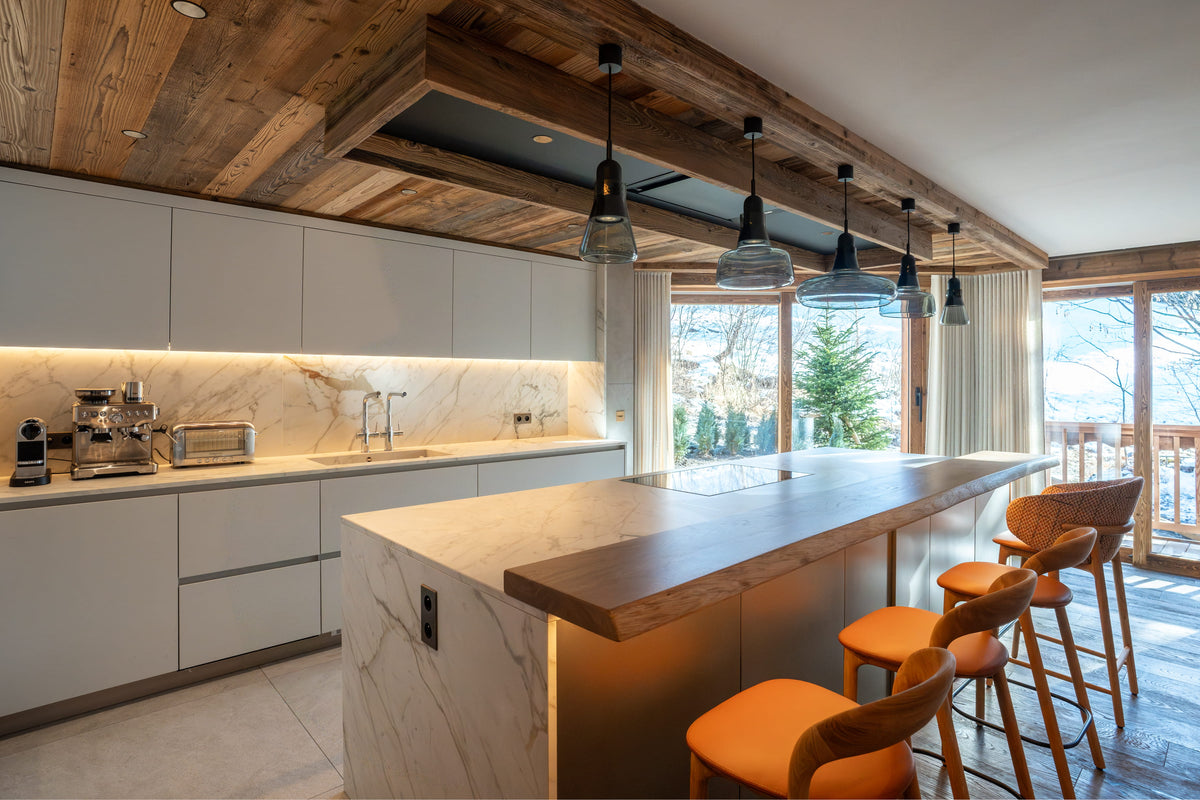 A modern kitchen interior with white cabinetry, a marble backsplash, a central island with a wood countertop, black pendant lights, and orange bar stools. Large windows and wooden ceiling beams are visible, with a snowy outdoor scene in the background.