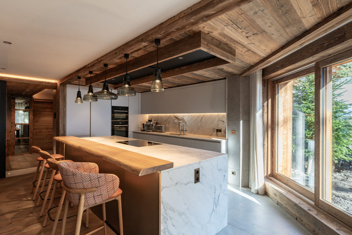 A modern kitchen island with a light wood countertop and white marble base, set in a rustic interior with exposed wooden ceiling beams, pendant lights, and bar stools. Large windows on the right show greenery outside.