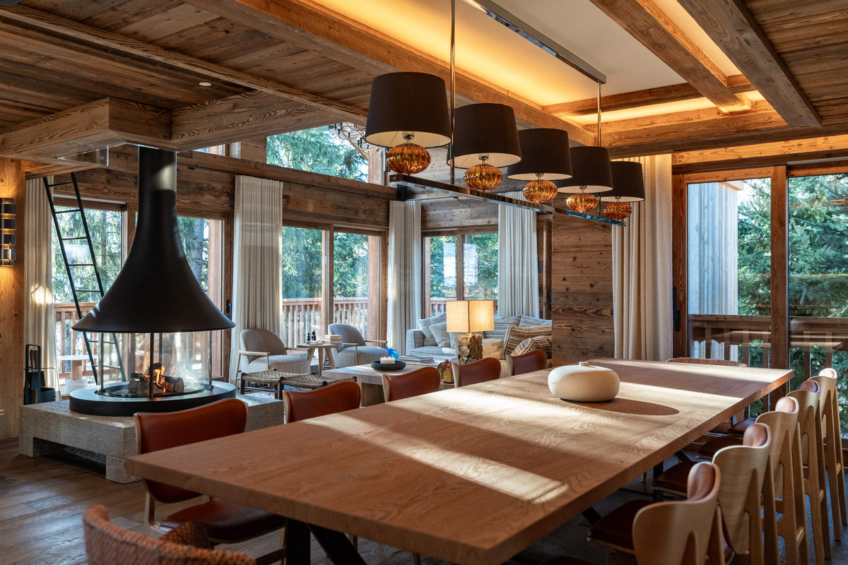 A large rustic wooden dining table with matching chairs in a cozy, sunlit chalet-style dining room featuring a fireplace, wood ceiling beams, and large windows.