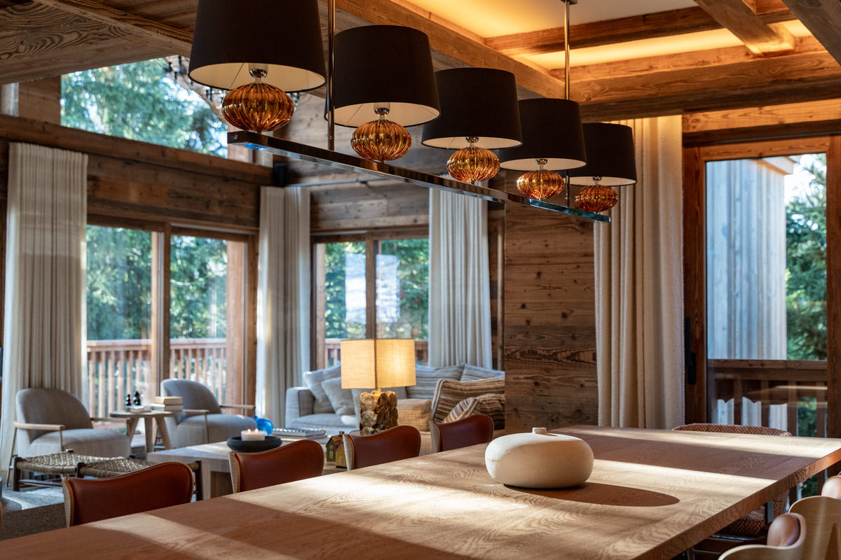 A modern dining room interior with a long wooden table in the foreground, a row of black drum pendant lights with amber glass bases hanging above, wood-paneled walls, large windows, and outdoor views.