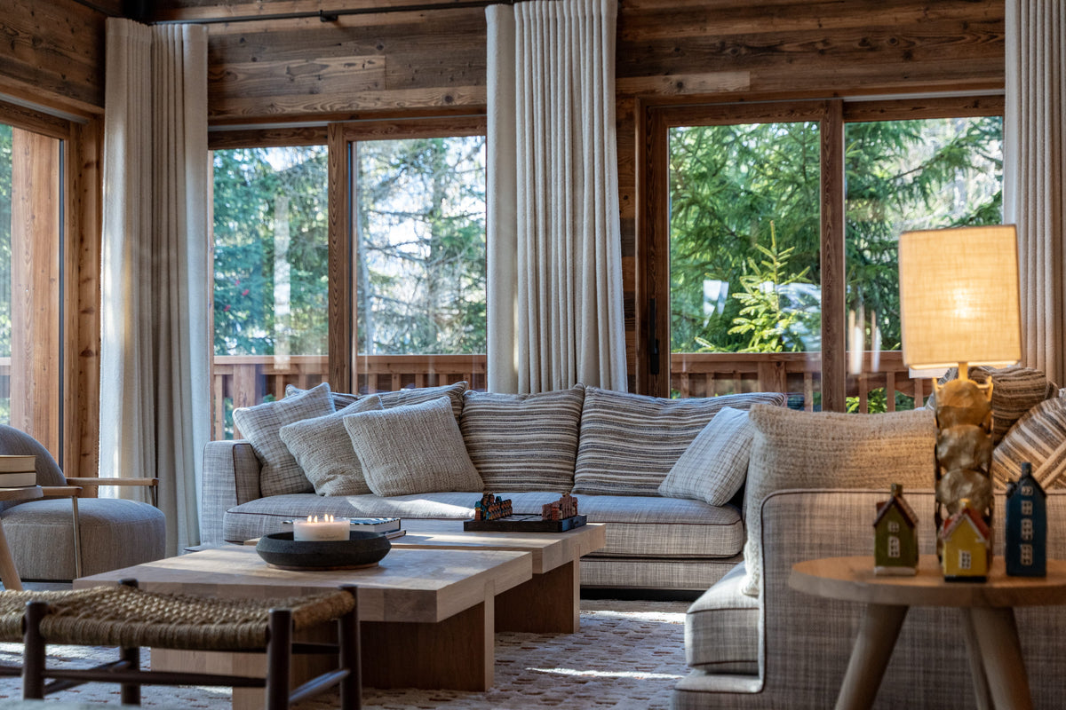 A cozy, rustic mountain chalet living room featuring a large L-shaped sectional sofa with striped beige and gray cushions, a low wooden coffee table, a woven ottoman, and a warm glowing table lamp by large windows with wood-paneled walls.