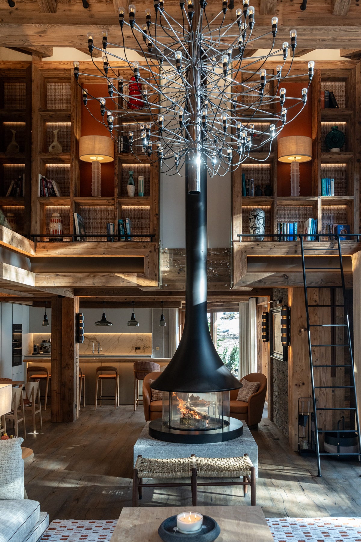 A large black central fireplace with a tall flue and a dramatic tree-style chandelier hanging from the ceiling in a rustic wooden loft living room.