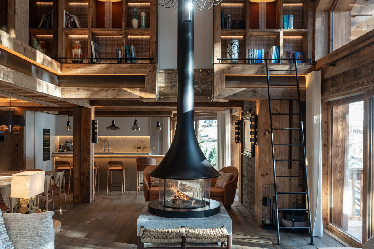 A rustic mountain chalet interior with a large black central fireplace chimney, exposed wood beams, loft shelves, a ladder, and a modern kitchen in the background.