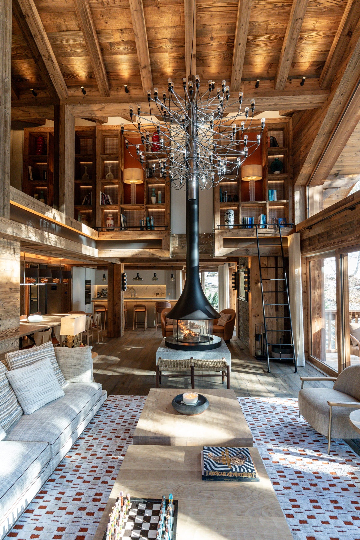A high-ceilinged rustic lodge interior with exposed wood beams, built-in shelving, a central black fireplace with a long flue, a large metal chandelier, a patterned tile floor, and multiple seating areas.