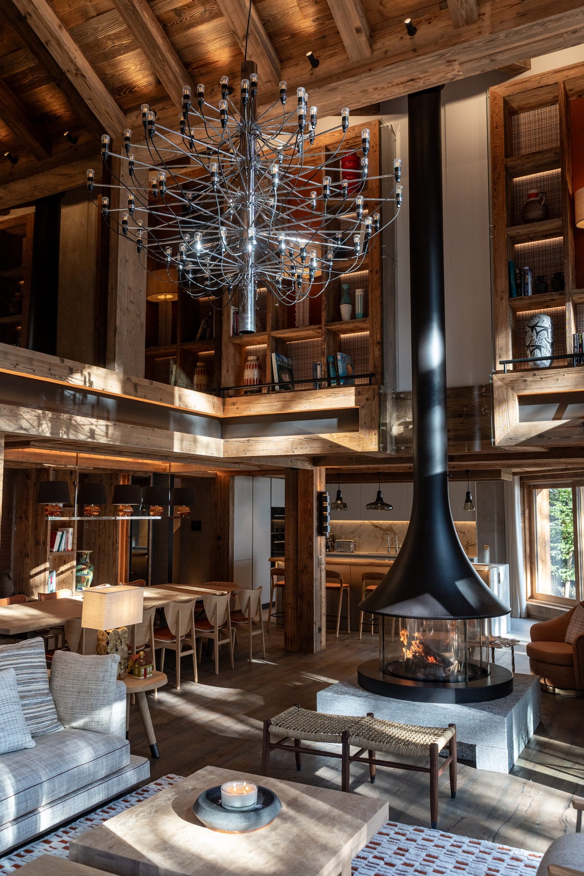 A rustic, high-ceilinged lodge-style living room with exposed wood beams, a large black spherical chandelier, built-in shelving, and a central black fireplace with a tall flue. The room includes seating, tables, and warm ambient lighting.