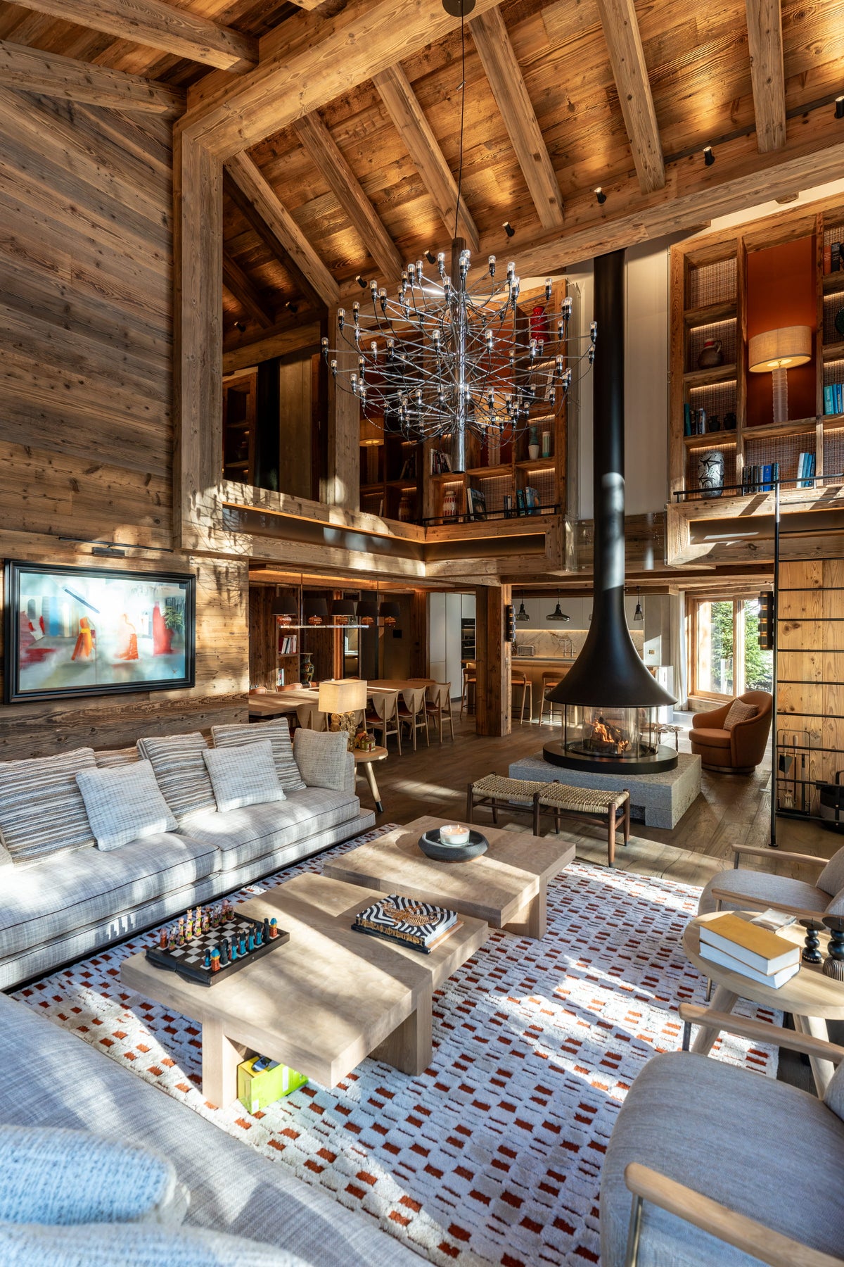A high-ceilinged rustic lodge living room with exposed wood beams, a large white sectional sofa, patterned area rugs, wooden coffee tables, built-in shelving, and a black central fireplace chimney.
