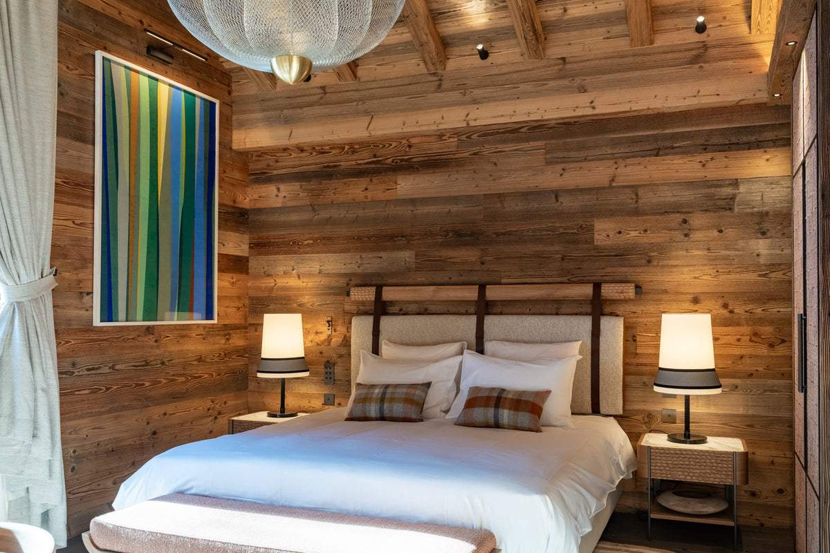 A rustic bedroom with wood-paneled walls and ceiling, featuring a neatly made bed with white linens, two bedside tables with lit lamps, a large colorful abstract wall art piece, and a hanging pendant light overhead.