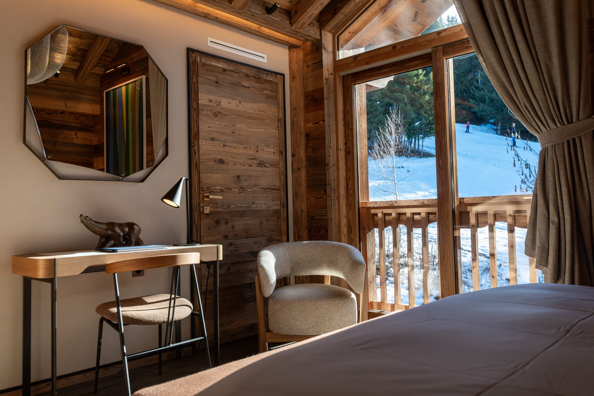 A cozy mountain chalet bedroom featuring a wooden desk with a matching chair, a large octagonal wall mirror, and a plush armchair. Large windows with brown curtains offer a view of a snowy outdoor landscape, and a bed is visible in the foreground.