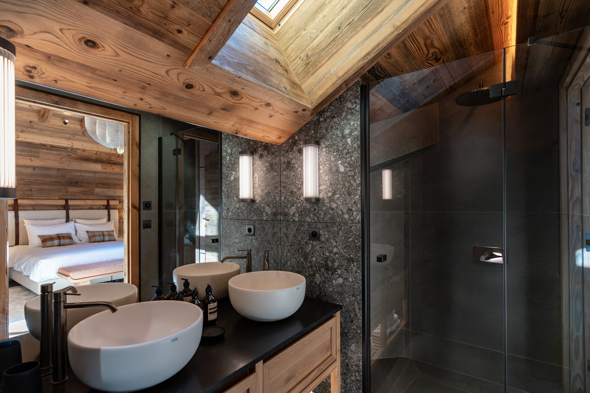 A luxury modern bathroom with a dark wood ceiling, stone walls, two white vessel sinks on a black countertop, and a glass shower enclosure.