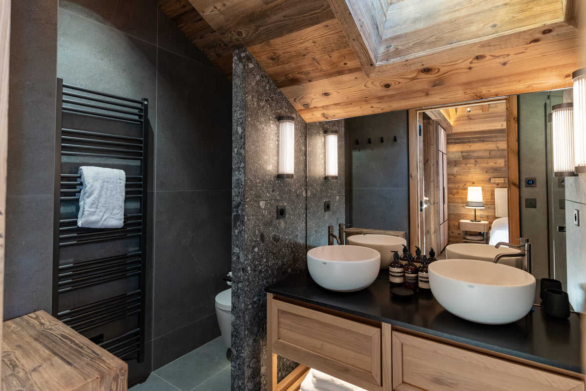A luxury modern bathroom featuring a double vanity with two white vessel sinks, a large wood and stone mirror, a dark stone wall, a black heated towel rail, and a rustic wooden ceiling.
