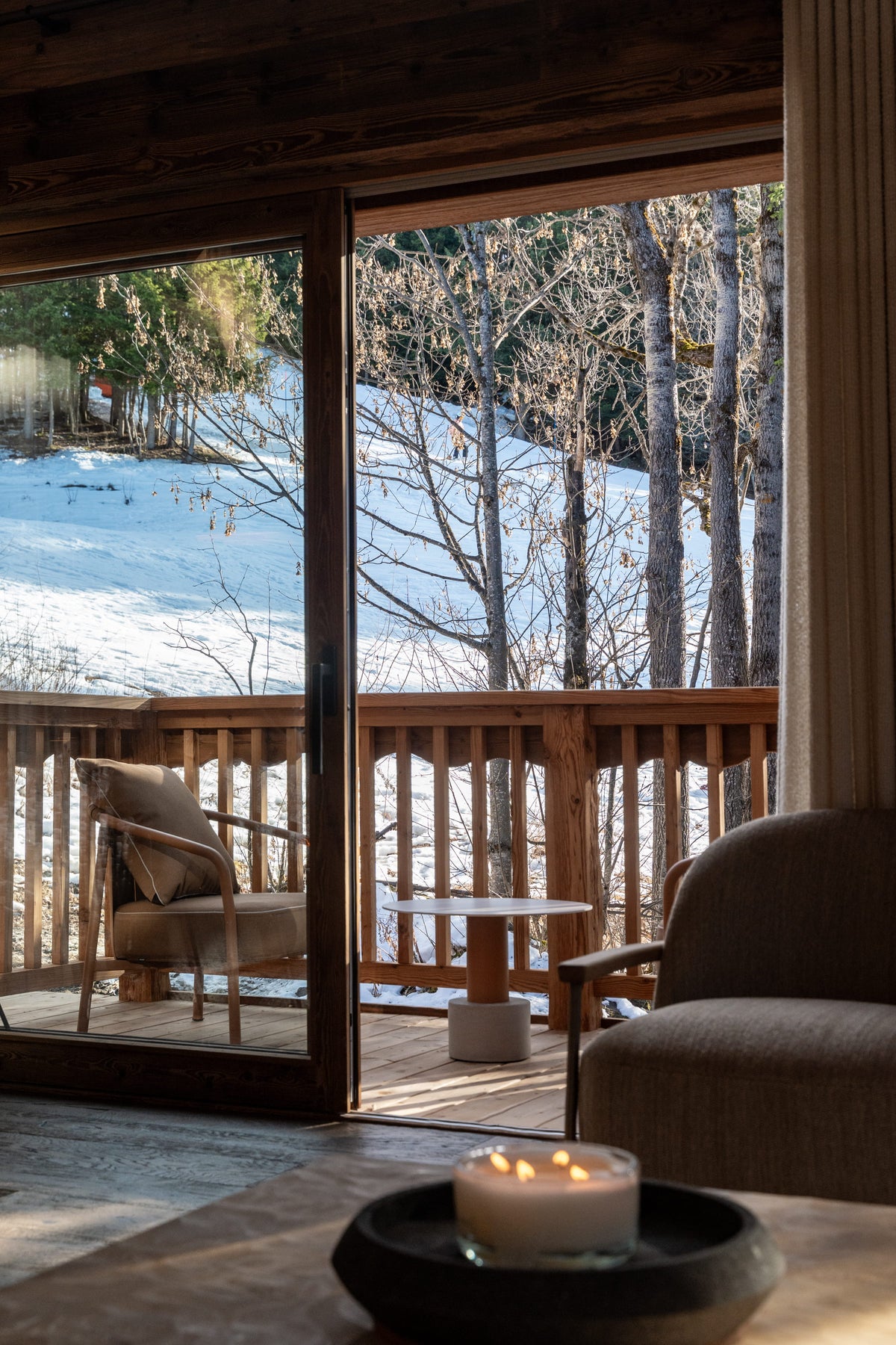 A cozy interior scene with large glass doors opening to a snowy outdoor deck and forest. A cushioned chair, side table, upholstered armchair, and a lit candle on a table are visible, creating a warm, rustic cabin atmosphere.
