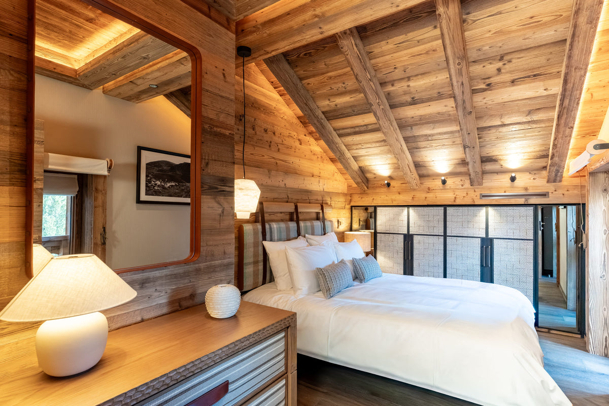 A rustic mountain-style bedroom with a large bed dressed in white linens, set against wood-paneled walls and a sloped wooden ceiling with exposed beams. A wooden dresser with a white table lamp is visible in the foreground, and a glass door is open in the background.