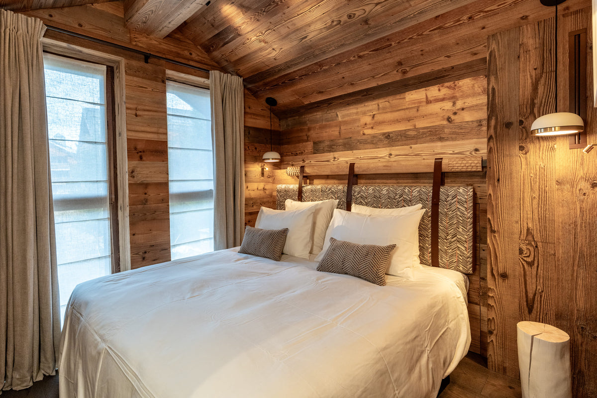 A cozy bedroom interior with a neatly made bed dressed in white linens and two taupe accent pillows, set against rustic wood-paneled walls and ceiling with large windows and warm wall sconces.