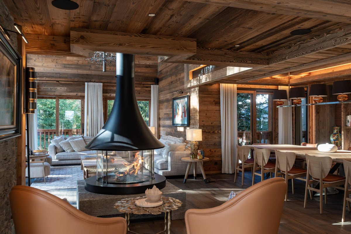 A luxurious rustic chalet interior featuring a large black cone-shaped fireplace with a glass firebox, surrounded by light wood walls and ceiling, beige leather armchairs, a dining area, and large windows looking out onto a forest.