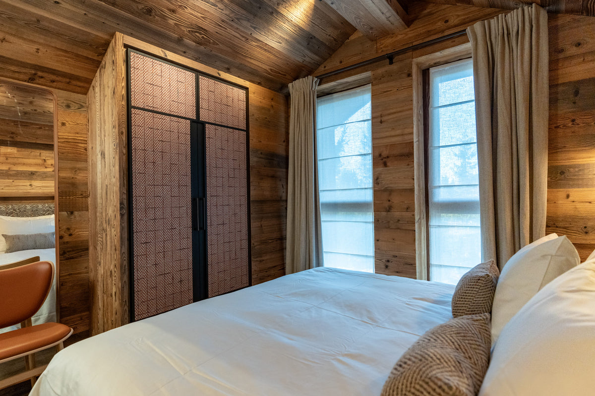 A cozy bedroom interior with a large bed dressed in white linens, wood-paneled walls and ceiling, two tall windows with beige curtains, and a tall built-in wardrobe with pinkish-brown doors and black framing.